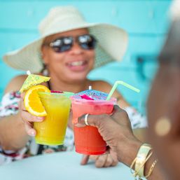 Two colorful tropical cocktails with paper umbrellas and an orange slice clinking on a sunny patio, smiling person in a wide sun hat and sunglasses blurred in the background.