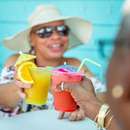 Two colorful tropical cocktails with paper umbrellas and an orange slice clinking on a sunny patio, smiling person in a wide sun hat and sunglasses blurred in the background.