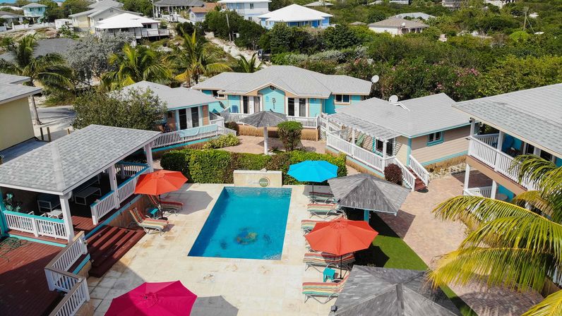Aerial view of a tropical beachside cottage courtyard with a rectangular swimming pool, colorful umbrellas and lounge chairs, palm trees and pastel bungalows — bright vacation rental vibe.