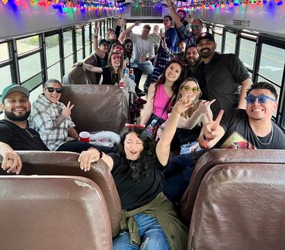 Party bus interior with colorful string lights and a large group of adults smiling, posing and holding drinks, many flashing peace signs in a lively celebration.