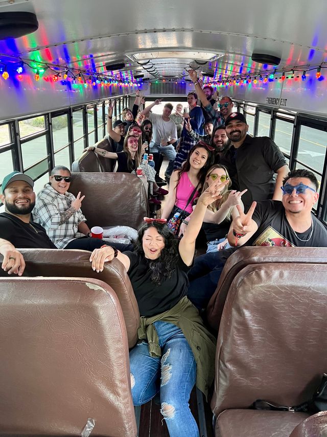 Party bus interior with colorful string lights and a large group of adults smiling, posing and holding drinks, many flashing peace signs in a lively celebration.