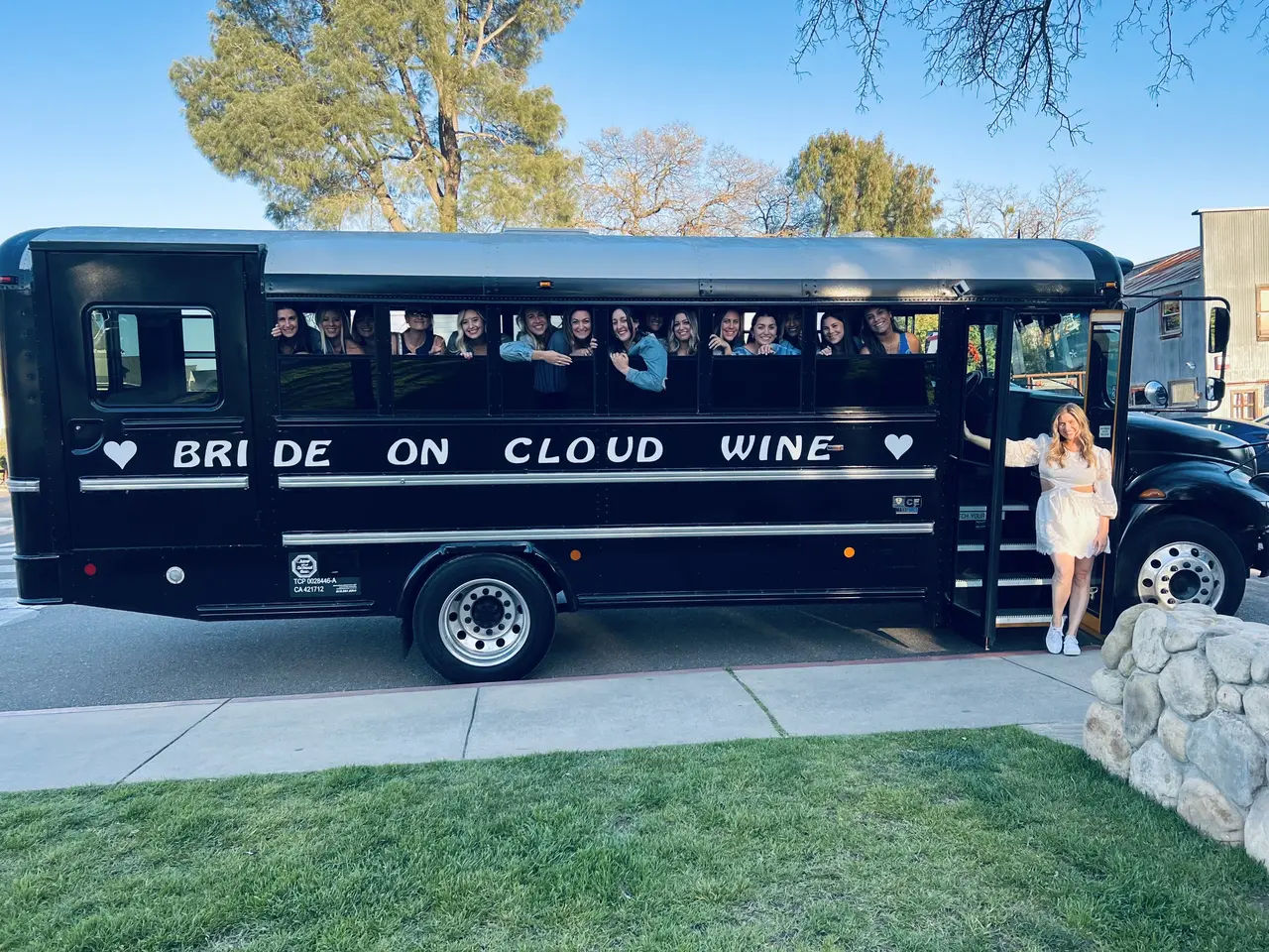 Black party bus reading 'BRIDE ON CLOUD WINE' parked on a tree-lined street, smiling bachelorette crew peeking from windows and the bride in a white dress standing at the open door under a clear blue sky.