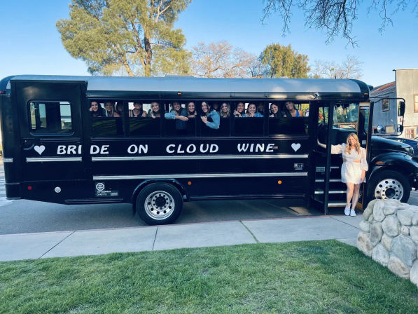 Black party bus reading 'BRIDE ON CLOUD WINE' parked on a tree-lined street, smiling bachelorette crew peeking from windows and the bride in a white dress standing at the open door under a clear blue sky.