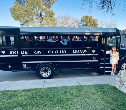 Black party bus reading 'BRIDE ON CLOUD WINE' parked on a tree-lined street, smiling bachelorette crew peeking from windows and the bride in a white dress standing at the open door under a clear blue sky.