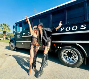 Two people in festival outfits—one in a sheer black dress with cowboy boots, the other in a black jacket, lace-up shorts and tall platform boots—posing with peace signs beside a black party bus on a sunny day with a palm tree in the background.