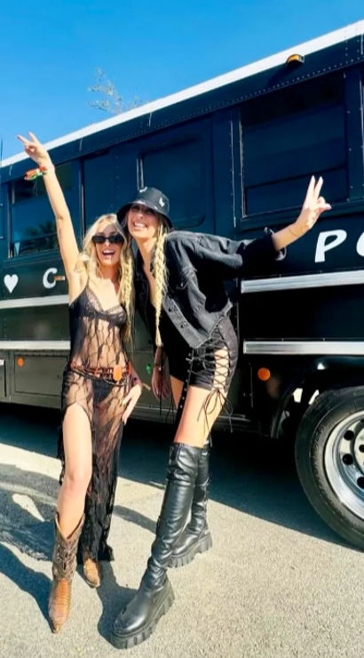 Two people in festival outfits—one in a sheer black dress with cowboy boots, the other in a black jacket, lace-up shorts and tall platform boots—posing with peace signs beside a black party bus on a sunny day with a palm tree in the background.