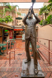Life-size bronze statue of a trumpeter with trumpet raised, standing on wet brick steps in an outdoor courtyard with green bistro tables and a neon Cajun food sign in the background.