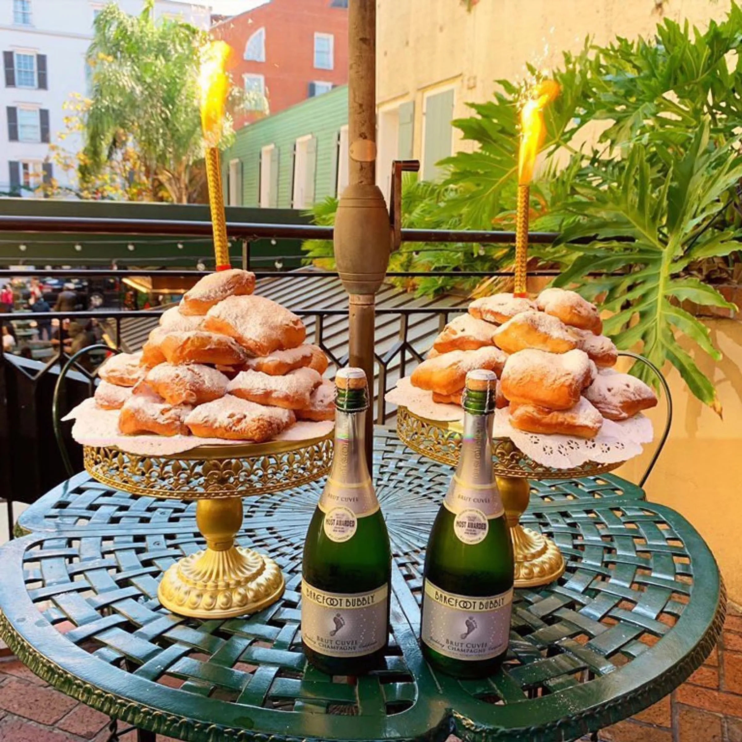 Two towers of powdered-sugar beignets on gold cake stands with sparklers and two mini champagne bottles on a green bistro balcony table — New Orleans–style outdoor brunch scene.