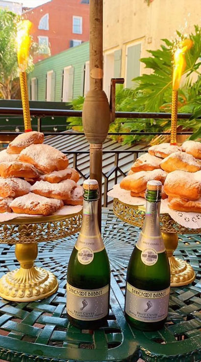 Two towers of powdered-sugar beignets on gold cake stands with sparklers and two mini champagne bottles on a green bistro balcony table — New Orleans–style outdoor brunch scene.