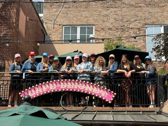 Smiling group of women in colorful trucker hats and denim jackets gathered on an outdoor balcony patio against an urban brick building, pink marquee sign on the railing.