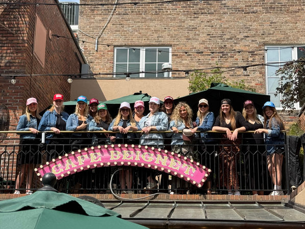 Smiling group of women in colorful trucker hats and denim jackets gathered on an outdoor balcony patio against an urban brick building, pink marquee sign on the railing.