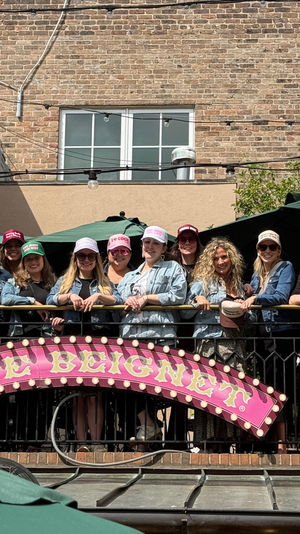 Smiling group of women in colorful trucker hats and denim jackets gathered on an outdoor balcony patio against an urban brick building, pink marquee sign on the railing.