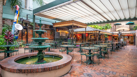 Covered brick courtyard cafe with a tiered green fountain in the foreground, wrought‑iron tables and chairs, a small stage with string lights and a beignet sign evoking a lively New Orleans French Quarter atmosphere.