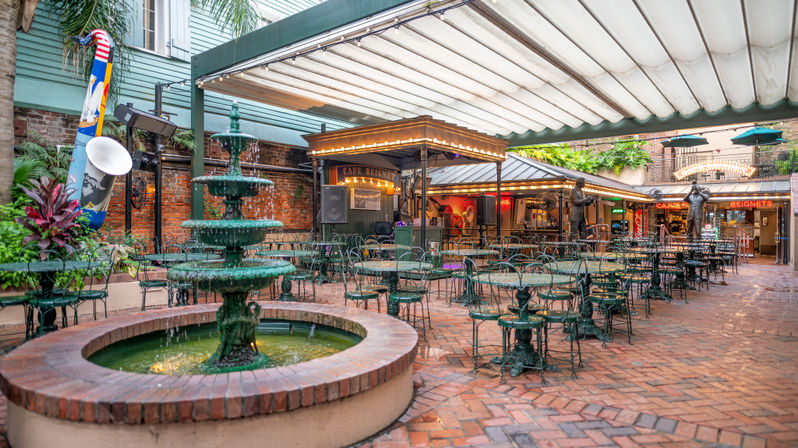 Covered brick courtyard cafe with a tiered green fountain in the foreground, wrought‑iron tables and chairs, a small stage with string lights and a beignet sign evoking a lively New Orleans French Quarter atmosphere.