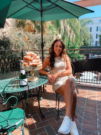 Smiling woman in a cream dress on a sunny outdoor patio brunch, seated at a green wrought-iron table with a tower of powdered donuts on a cake stand and a bottle of sparkling wine, palm trees and terrace buildings in the background.