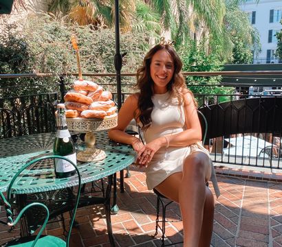 Smiling woman in a cream dress on a sunny outdoor patio brunch, seated at a green wrought-iron table with a tower of powdered donuts on a cake stand and a bottle of sparkling wine, palm trees and terrace buildings in the background.