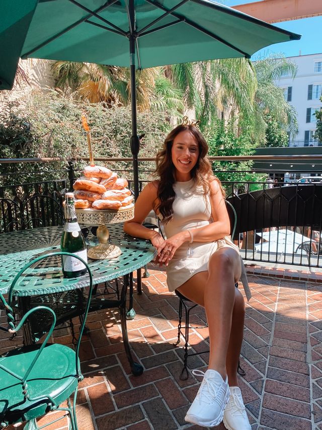 Smiling woman in a cream dress on a sunny outdoor patio brunch, seated at a green wrought-iron table with a tower of powdered donuts on a cake stand and a bottle of sparkling wine, palm trees and terrace buildings in the background.