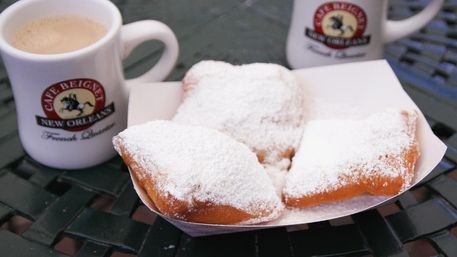 Three powdered New Orleans-style beignets in a paper tray beside two coffee mugs on a green metal cafe table.