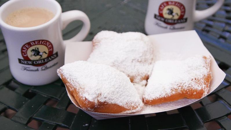 Three powdered New Orleans-style beignets in a paper tray beside two coffee mugs on a green metal cafe table.