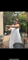 Smiling woman in a white sundress and sunglasses popping a champagne bottle on a brick patio surrounded by potted greenery, metal bistro chairs and an ice bucket.