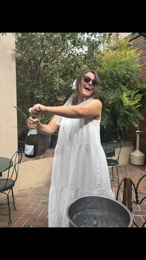 Smiling woman in a white sundress and sunglasses popping a champagne bottle on a brick patio surrounded by potted greenery, metal bistro chairs and an ice bucket.