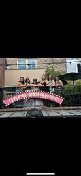 Six women smiling on a second-floor outdoor balcony above a pink illuminated marquee cafe sign, with brick building facade and patio umbrellas in the background