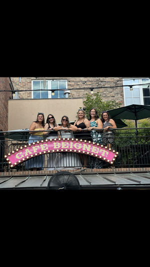 Six women smiling on a second-floor outdoor balcony above a pink illuminated marquee cafe sign, with brick building facade and patio umbrellas in the background