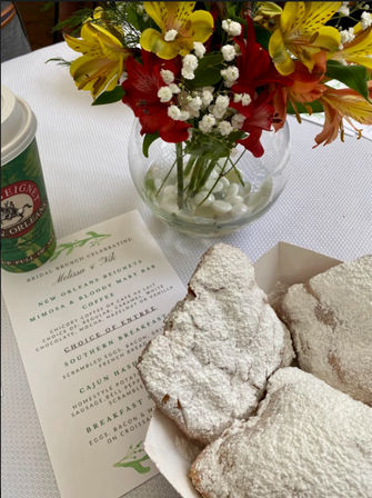 New Orleans-style powdered beignets in a paper tray beside a coffee cup, bright yellow and red floral centerpiece in a glass vase, and a printed brunch menu on a white tablecloth.