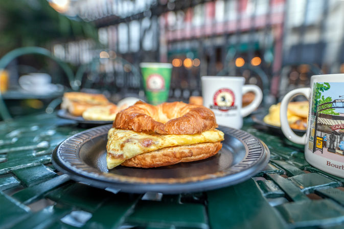 Crispy croissant breakfast sandwich with folded egg on a black plate, surrounded by coffee mugs on a green outdoor bistro table with warm bokeh lights in the background.