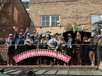 Group of women in denim jackets and colorful caps cheering on an outdoor balcony of an urban brick building, string lights overhead and a pink marquee sign below on a sunny rooftop patio.