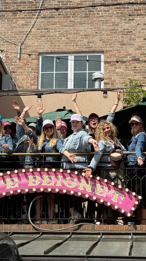 Group of women in denim jackets and colorful caps cheering on an outdoor balcony of an urban brick building, string lights overhead and a pink marquee sign below on a sunny rooftop patio.