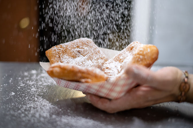 New Orleans-style beignets dusted with powdered sugar falling into a checkered paper tray held in hands