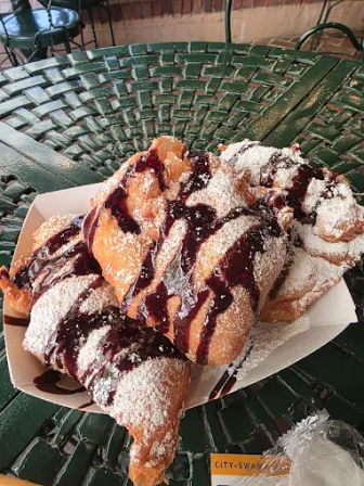 Paper tray of fried beignet-style pastries dusted with powdered sugar and drizzled with chocolate sauce on a green metal outdoor bistro table at a cafe