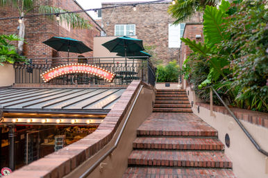 Cozy urban courtyard with brick steps leading to a terrace café — green patio umbrellas, string lights, lush tropical plants and a glowing marquee over a glass-roofed entrance.