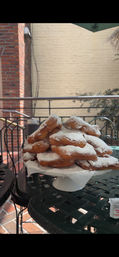 Stack of fresh powdered-sugar beignets on a white cake stand at an outdoor café patio with a wrought-iron table and brick wall backdrop