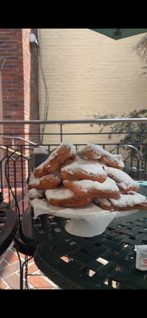 Stack of fresh powdered-sugar beignets on a white cake stand at an outdoor café patio with a wrought-iron table and brick wall backdrop