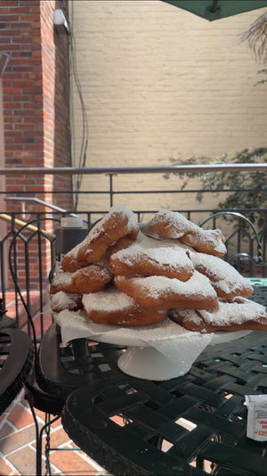 Stack of fresh powdered-sugar beignets on a white cake stand at an outdoor café patio with a wrought-iron table and brick wall backdrop