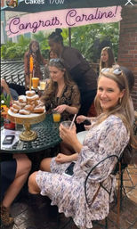 Outdoor patio brunch with smiling women around a wrought-iron table, a gold cake stand piled with powdered pastries, glasses of orange mimosas, and a pink celebration banner overhead.