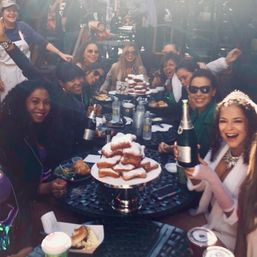Group of women enjoying an outdoor patio brunch party, smiling around a table with a tower of powdered-sugar beignets, pastries, coffee cups and champagne bottles.
