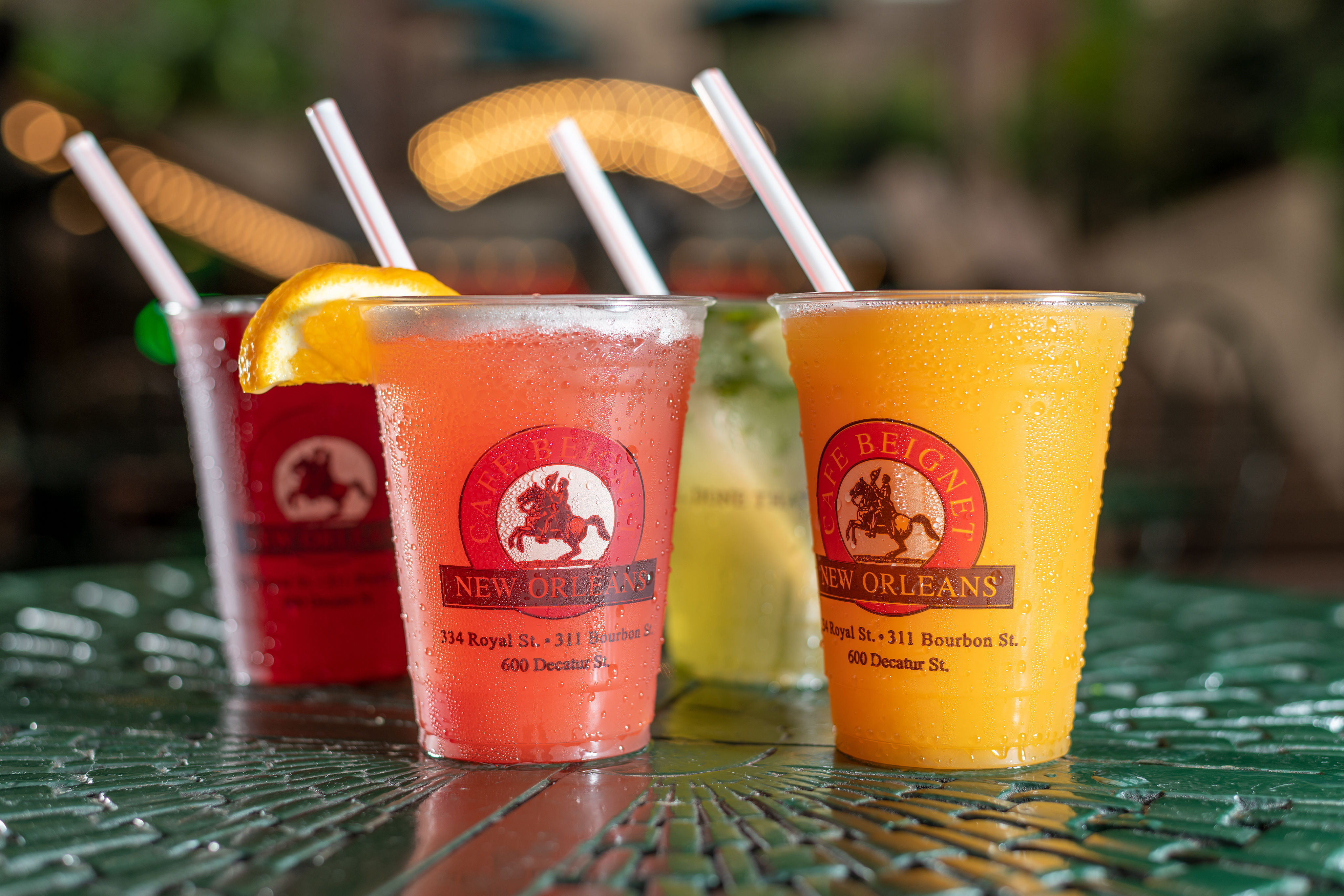 Four colorful frozen cocktails in plastic cups with straws on a wet green metal patio table, bright bokeh lights and blurred outdoor cafe background evoking New Orleans street-side summer vibes.