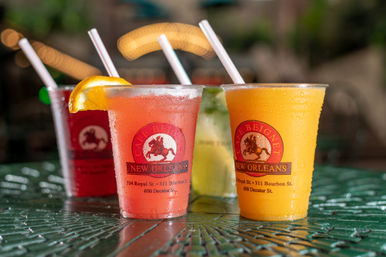 Four colorful frozen cocktails in plastic cups with straws on a wet green metal patio table, bright bokeh lights and blurred outdoor cafe background evoking New Orleans street-side summer vibes.