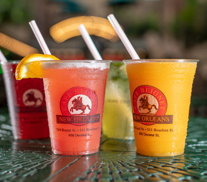 Four colorful frozen cocktails in plastic cups with straws on a wet green metal patio table, bright bokeh lights and blurred outdoor cafe background evoking New Orleans street-side summer vibes.