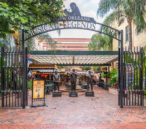 New Orleans courtyard under a wrought-iron arch reading 'Musical Legends Park' featuring three bronze jazz musician statues, palm trees, brick walkway and outdoor café seating.