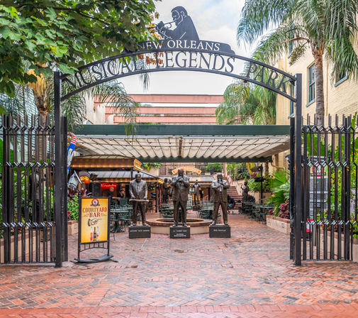 New Orleans courtyard under a wrought-iron arch reading 'Musical Legends Park' featuring three bronze jazz musician statues, palm trees, brick walkway and outdoor café seating.