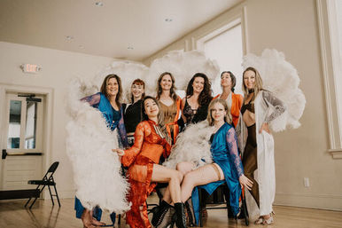 Eight burlesque performers in colorful robes and lingerie pose with large white feather fans in a sunlit rehearsal studio with wood floors and tall windows.