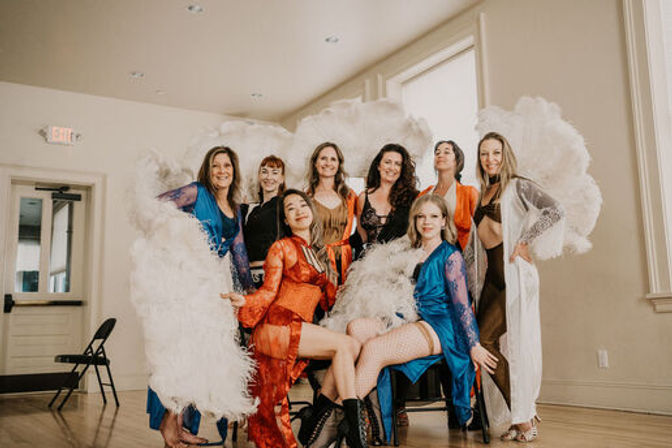 Eight burlesque performers in colorful robes and lingerie pose with large white feather fans in a sunlit rehearsal studio with wood floors and tall windows.