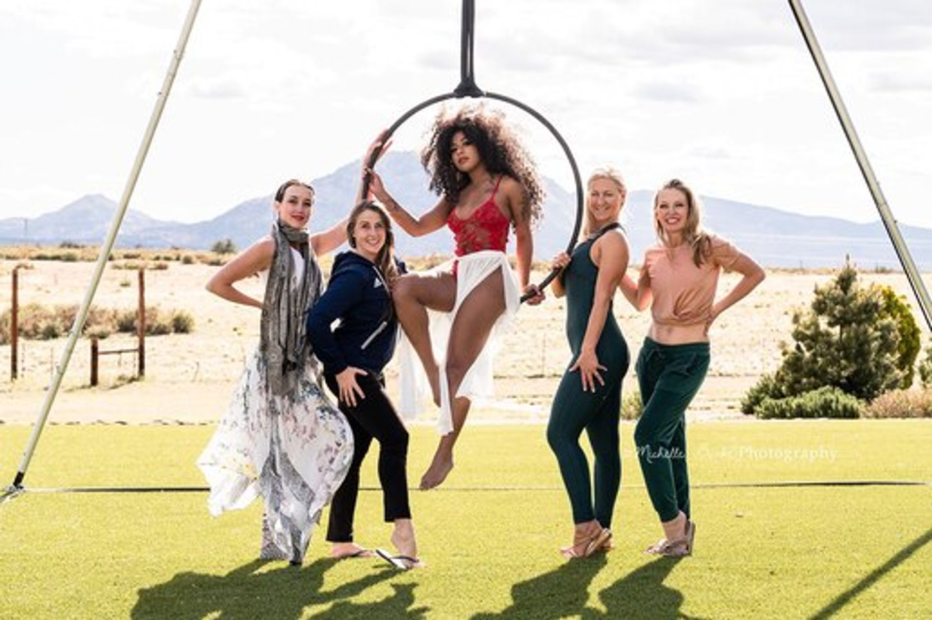 Five women at an outdoor aerial hoop photoshoot: one seated in a suspended hoop wearing a red top and white skirt, four others smiling on turf against desert and mountain scenery under a sunny sky.