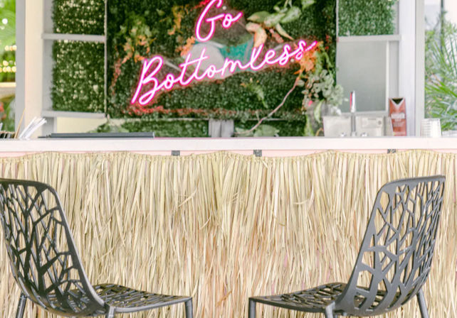 Tropical-chic outdoor bar with straw tiki front, two black metal lattice chairs, lush green plant wall and pink neon 'Go Bottomless' sign