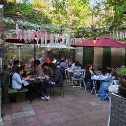Cozy urban garden patio courtyard with hanging greenery and string lights, red umbrellas and tables of people enjoying al fresco dining.