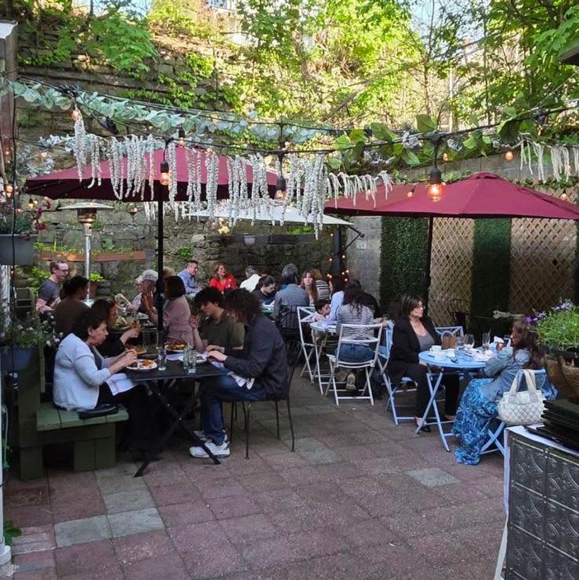 Cozy urban garden patio courtyard with hanging greenery and string lights, red umbrellas and tables of people enjoying al fresco dining.